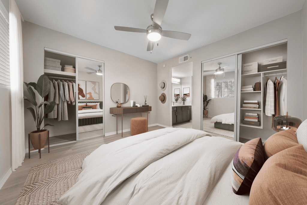 Modern bedroom with neutral decor, a neatly made bed, cozy pillows, mirrored closet doors, open wardrobe, wooden vanity with round mirror, plants, and a ceiling fan. Natural light fills the room from large windows.