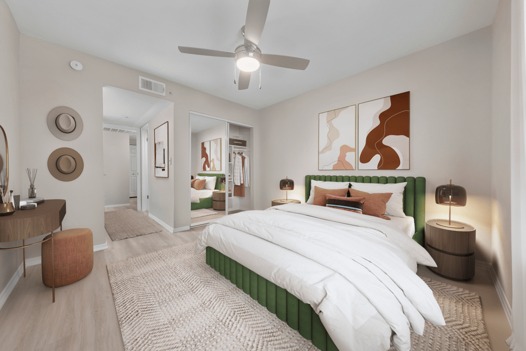 Modern bedroom with a green upholstered bed, white bedding, two abstract art pieces above the headboard, nightstands with lamps, a ceiling fan, wall-mounted hats, and a mirror reflecting the room.