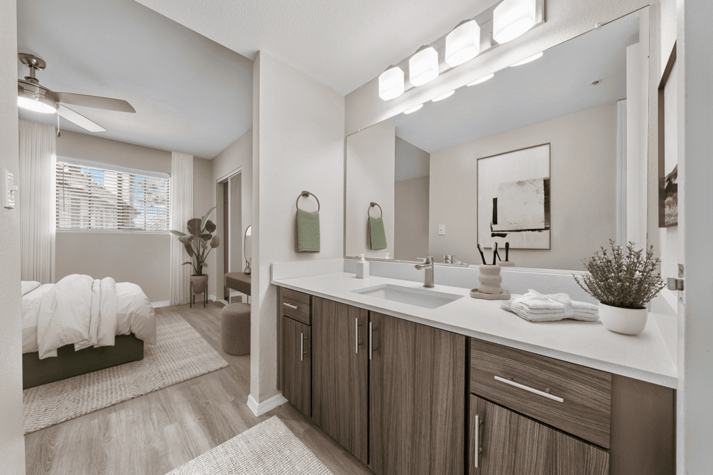 Modern bathroom with wood-finish cabinets, a large mirror, and bright lighting, opening into a bedroom with a made bed, large window, ceiling fan, and neutral decor. A potted plant sits on the vanity.