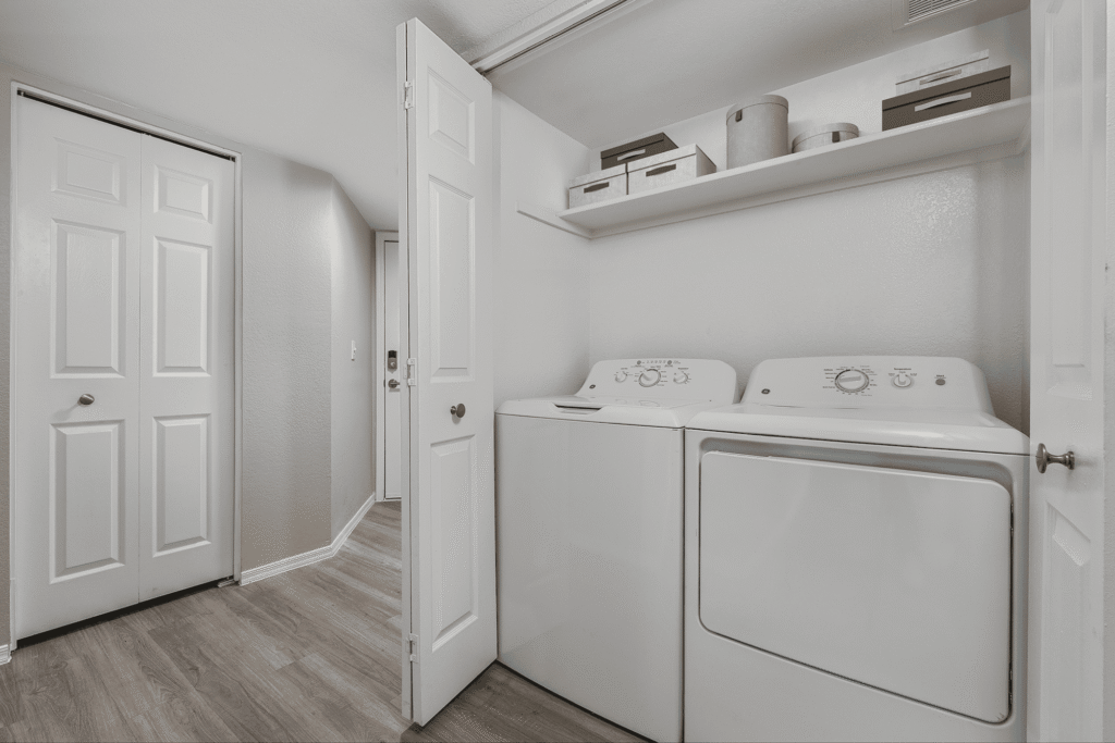 A laundry nook with a washing machine and dryer side by side, white walls, wooden floor, and shelves above holding storage boxes and containers; the area is enclosed by white double doors.