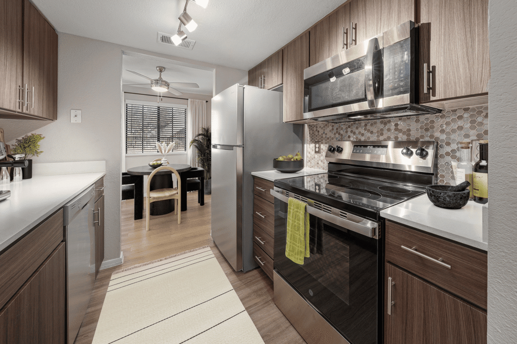 Modern kitchen with stainless steel appliances, wood cabinets, white countertops, and a hexagon tile backsplash. A dining area with a round table and four chairs is visible through a doorway. A beige rug runs along the kitchen floor.