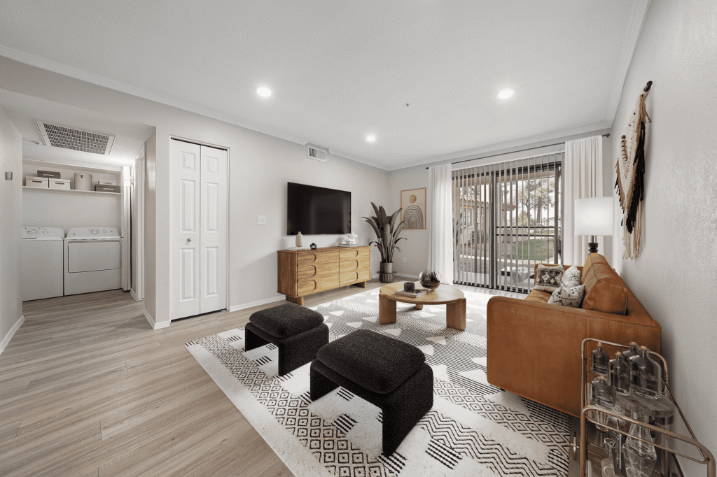 Modern living room with a tan sofa, patterned rug, two black ottomans, wooden coffee table, TV on a wooden console, large window with blinds, and a view of a laundry area with washer and dryer in the background.