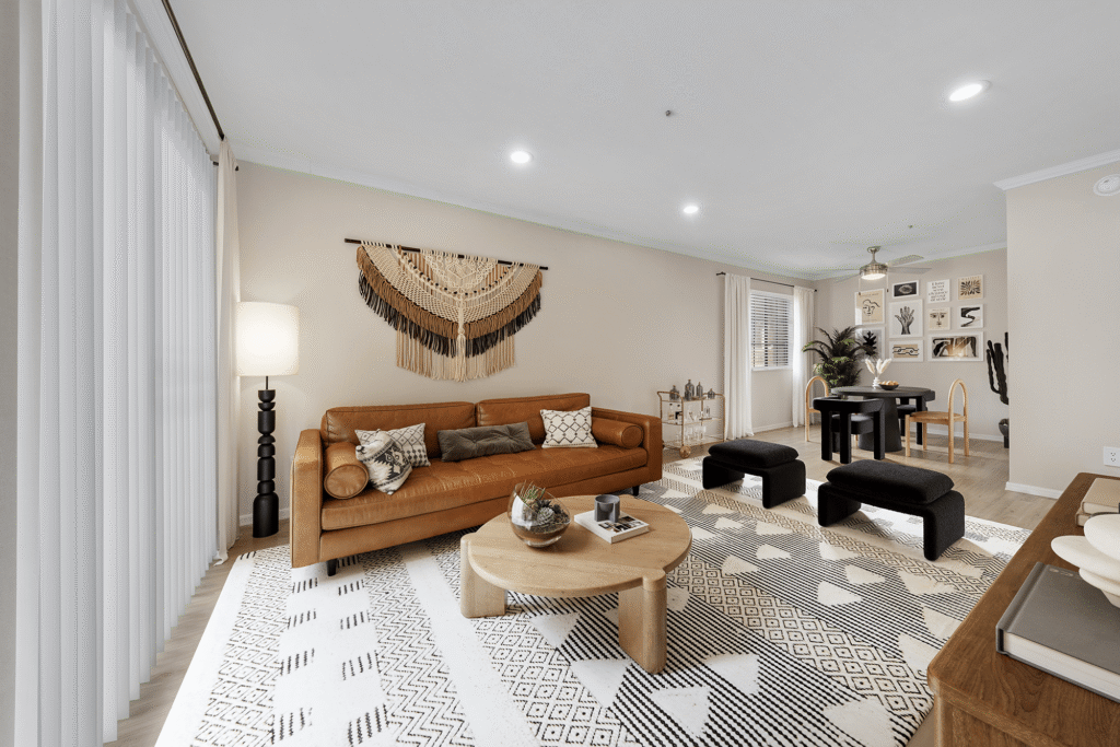A modern living room with a brown leather sofa, black-and-white geometric rug, round wooden coffee table, black ottomans, wall art, and a dining area in the background with a gallery wall and dining table.