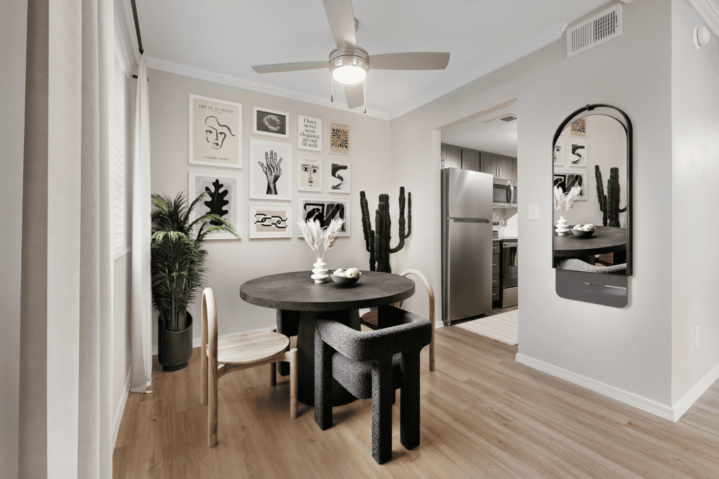 A modern dining area with a round black table, mixed-style chairs, wall art, potted plants, a ceiling fan, wood flooring, and a view into a kitchen with stainless steel appliances.