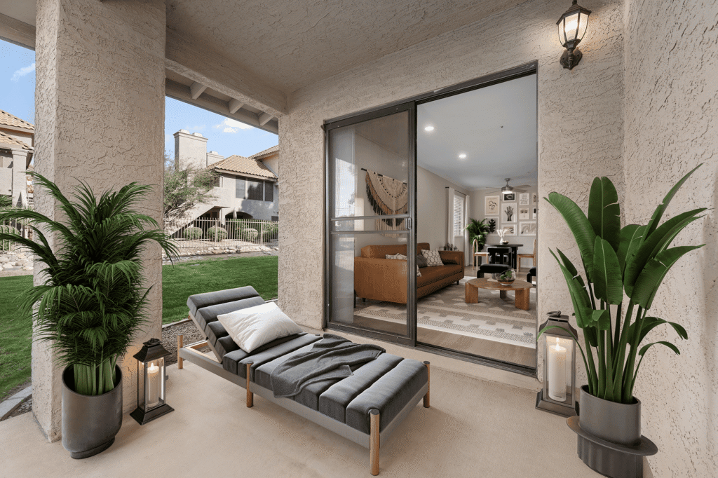 A covered patio with a cushioned lounge chair, two large potted plants, and lanterns, overlooking a grassy area. Nearby, glass doors lead to a cozy living room with modern decor.