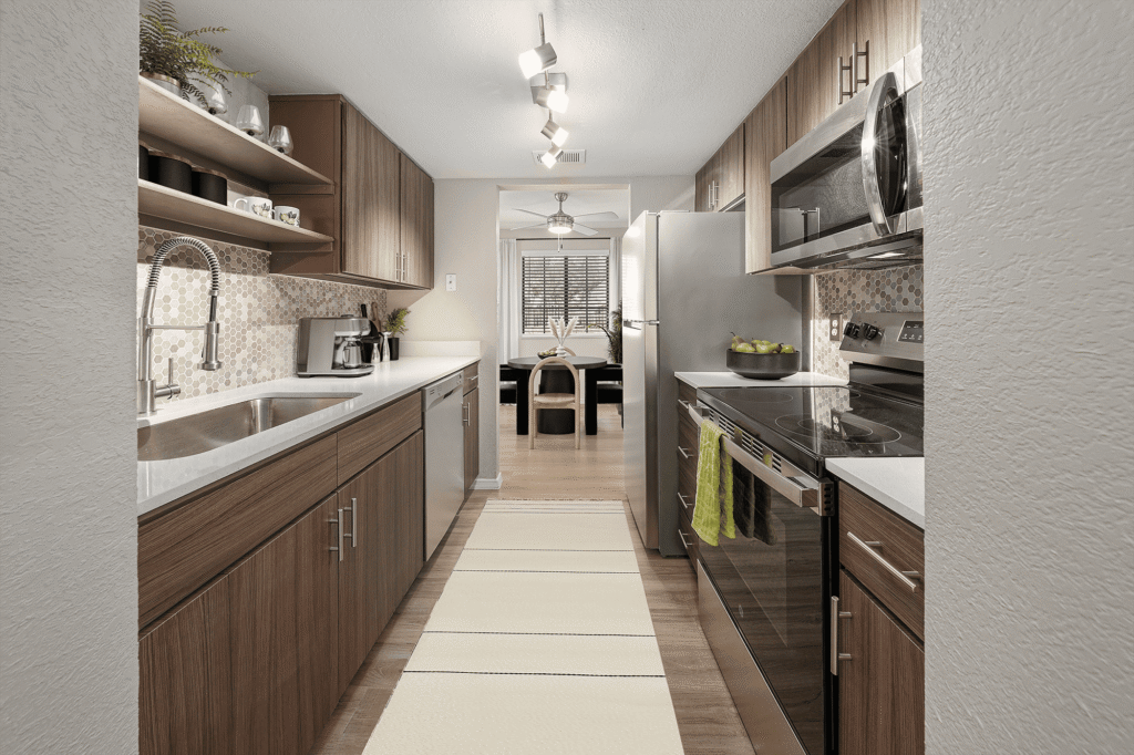 Modern galley kitchen with wood cabinets, stainless steel appliances, mosaic tile backsplash, and white countertops. A runner rug lines the floor, and a dining area with a window is visible at the far end.